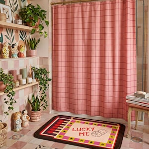 May include: A rectangular bath mat featuring a graphic design of a matchbox with the text LUCKY ME and an illustration of two dice. The mat has a cream center, a pink and yellow border with black stars, and a dark brown edge on a tiled floor.