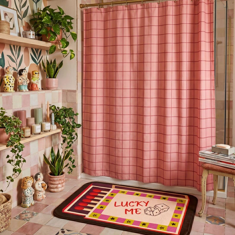 May include: A rectangular bath mat featuring a graphic design of a matchbox with the text LUCKY ME and an illustration of two dice. The mat has a cream center, a pink and yellow border with black stars, and a dark brown edge on a tiled floor.