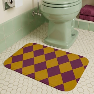 May include: A rectangular bath mat featuring a repeating diamond pattern in alternating shades of deep purple and mustard yellow. The mat has rounded corners and a flat, uniform texture, positioned on a white hexagonal tiled bathroom floor.