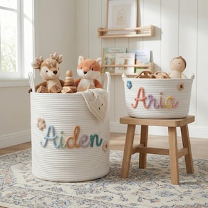 May include: Two white rope storage baskets with handles. The larger basket features the name Aiden in pastel yarn letters with small flower accents. The smaller basket, resting on a wooden stool, features the name Aria in pastel yarn letters.