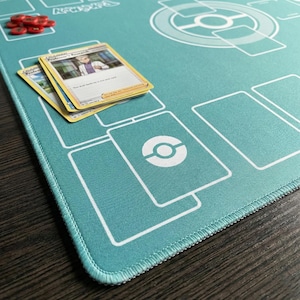 May include: Teal playmat with white outlines and a Pok&eacute;mon theme. Two trading cards and red tokens are on the mat. The mat has a stitched edge and a circular design with a Pok&eacute; Ball symbol.