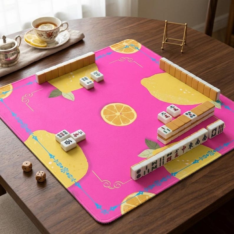 May include: A pink Mahjong game mat with a lemon design. The mat has a border with yellow and blue accents. Mahjong tiles and dice are arranged on the mat, ready for play. A cup of tea and a small metal tea pot are in the background.