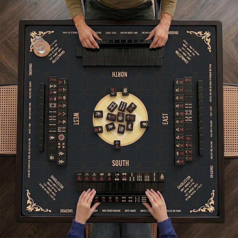 May include: A square mahjong game table featuring a dark surface with gold printed compass directions, Charleston instructions, and decorative corner flourishes. Black tiles with red and gold symbols are arranged in rows and a central cluster.