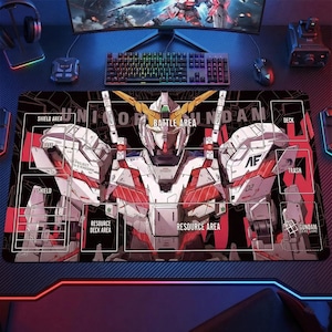 May include: Black gaming mousepad featuring a detailed illustration of a Gundam robot with red, white, and gold accents. The mat includes labeled areas for card game play, such as "Battle Area" and "Resource Area."