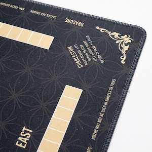 May include: A black Mahjong mat featuring gold printed text and grid patterns. The mat includes labels for East, Dragons, and Charleston rules, with decorative gold flourishes in the corner and stitched edges for a durable finish.