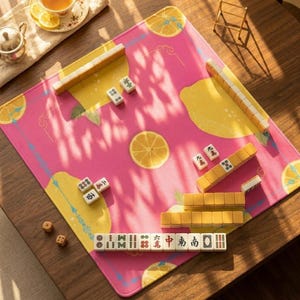 May include: A pink Mahjong game mat with yellow lemon slice designs and a border. The mat is set up with yellow and white tiles, dice, and a small wooden stand. A cup of tea with lemon is in the background.