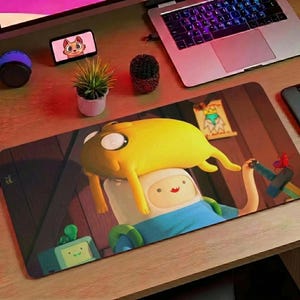May include: A rectangular mousepad featuring a colorful illustration of characters from the animated series Adventure Time. The design includes Finn and Jake in a whimsical scene, with a wooden background.
