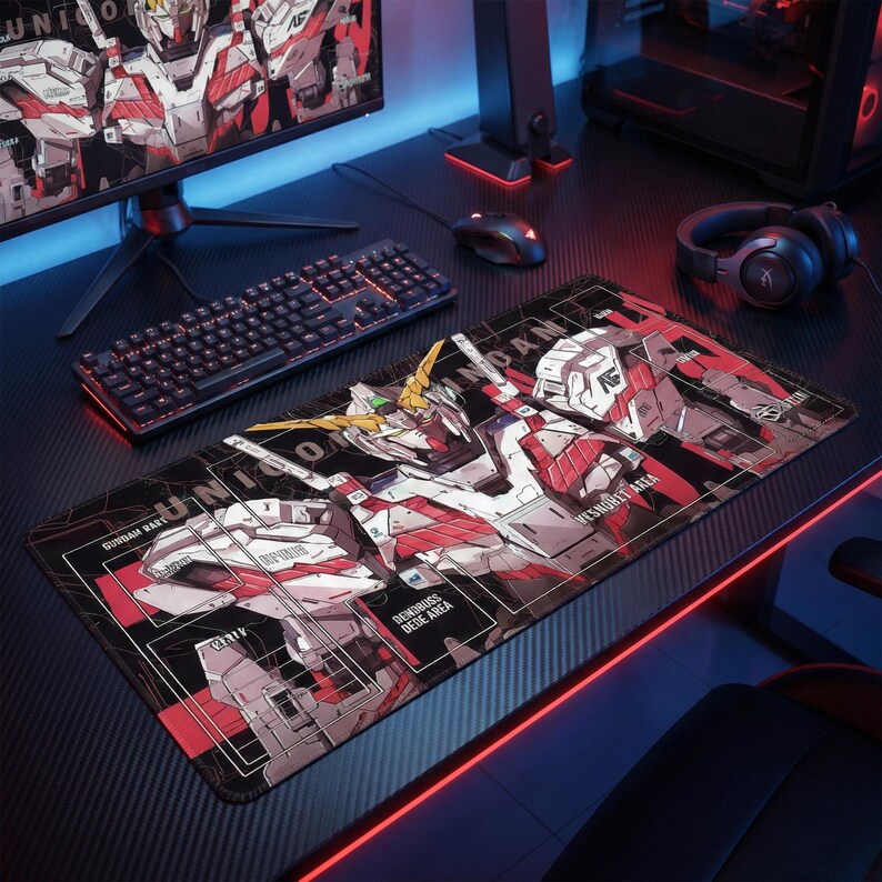 May include: A black and red rectangular gaming mousepad featuring a Gundam robot design. The mousepad has a black border and the text "UNICORN" is visible. The design includes red, white, and yellow accents. A keyboard, mouse, and monitor are also visible.