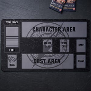 Peut inclure: Tapis de jeu de cartes One Piece noir et gris. Le tapis présente des zones étiquetées pour la Vie, le Personnage, le Coût et le Deck, avec un motif de carte du monde. Le texte "ONE PIECE CARD GAME" est en haut à gauche. Deux paquets de cartes sont visibles.