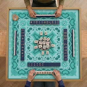 May include: A top-down view of a Mahjong game in progress. The teal game board features a decorative pattern and is surrounded by a wooden frame. Players are arranging tiles, with some tiles in a central pile.