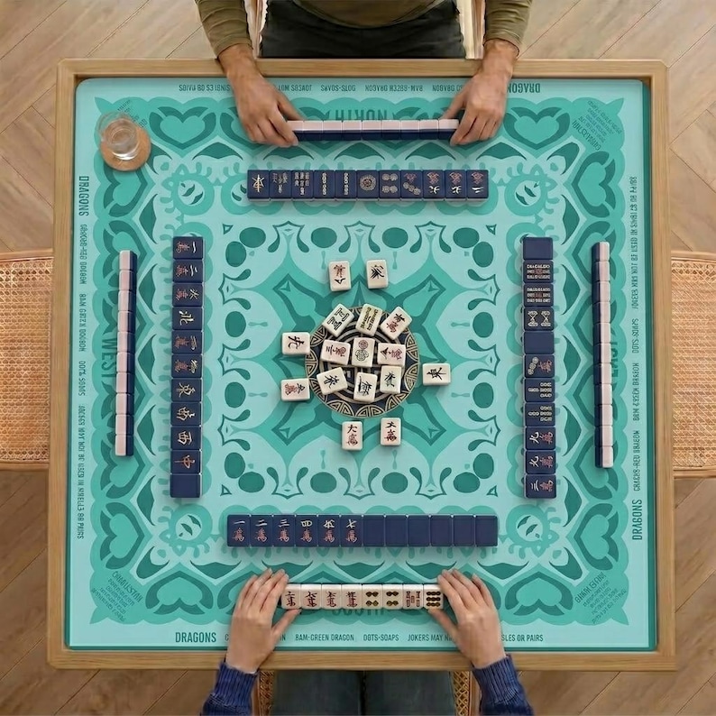 May include: A top-down view of a Mahjong game in progress. The teal game board features a decorative pattern and is surrounded by a wooden frame. Players are arranging tiles, with some tiles in a central pile.
