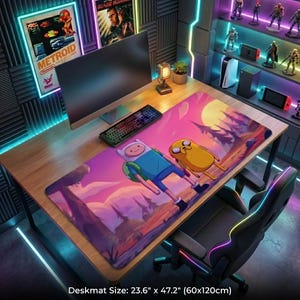 May include: A large desk mat featuring Finn and Jake from Adventure Time, set against a sunset landscape. The mat measures 23.6 inches by 47.2 inches. A keyboard and computer monitor are visible on the wooden desk, along with gaming accessories.