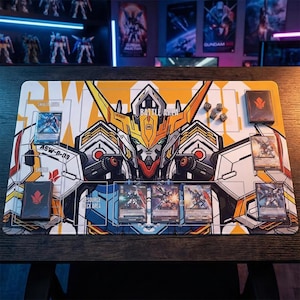 May include: A rectangular Gundam-themed playmat with a detailed illustration of a robot's face in yellow, orange, white, and black. The mat features designated areas for cards, dice, and a deck, with the words "BATTLE AREA" and "RESOURCE DECK AREA".