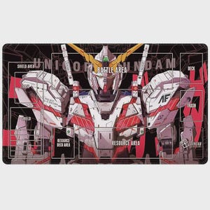 May include: A black and red playmat featuring a detailed illustration of a Gundam robot. The mat includes labeled areas for gameplay, such as "Battle Area," "Deck," and "Resource Area." The robot is white, red, and gold, with the text "UNICORN GUNDAM" across the top.