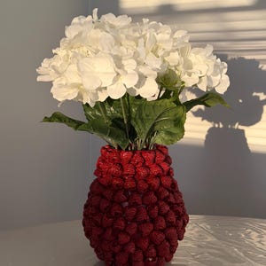 May include: A decorative vase with a textured red exterior, filled with white flowers and green leaves. The vase has a unique design, and the flowers are in full bloom, creating a striking contrast.