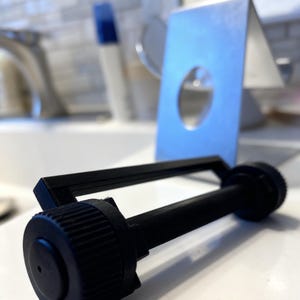 Puede incluir: Un objeto cilíndrico negro con una tapa de extremo circular y un cuerpo estriado, posiblemente un accesorio de baño. Un soporte de metal plateado con un recorte circular está en el fondo.