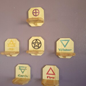 May include: A collection of wooden shelves, each featuring a symbol representing an element: Air, Earth, Fire, Water, and a pentagram. The shelves are made of light-coloured wood and mounted on a purple wall. Each shelf has a small ledge.