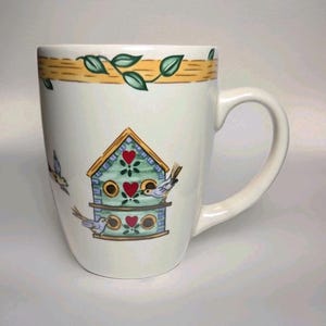 May include: White ceramic mug with a birdhouse design. The mug features a birdhouse with red hearts, blue accents, and birds. Green leaves and a yellow band with brown lines decorate the top rim. The mug has a curved handle.