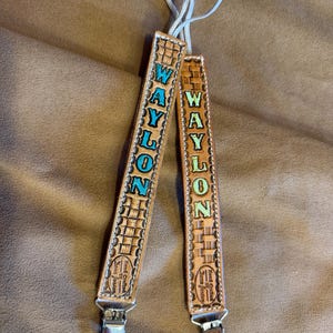 May include: Two tan leather pacifier clips featuring a basketweave pattern and the name WAYLON embossed in vertical lettering. The left clip has bright blue letters, while the right clip has light green letters. Each includes a metal clip and loop.