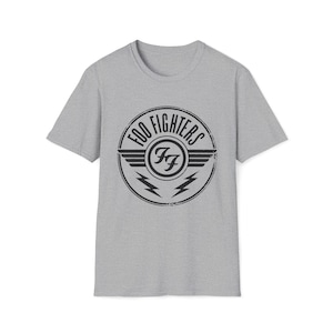 May include: A gray t-shirt featuring a black circular logo with the text "FOO FIGHTERS" and the letters "FF" inside. The logo includes wings and lightning bolts. The shirt is made of a soft material.