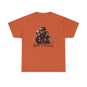 May include: Orange t-shirt featuring a sepia-toned graphic of the band Alice in Chains. The graphic includes band members, a microphone, and the band's name in a stylized font. The shirt is made of a soft material.