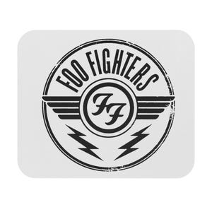 May include: White, rounded-corner square mousepad featuring the Foo Fighters logo. The logo is a black circular design with the band's name, wings, lightning bolts, and the letters "FF" in the center.