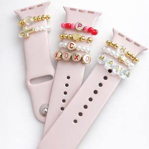 Può includere: Tre cinturini rosa con braccialetti di perline. Un cinturino ha un ciondolo a forma di croce dorata, un altro ha perline con lettere che compongono "XOXO", e il terzo ha "MAMA". I braccialetti presentano perline dorate, bianche e rosse.