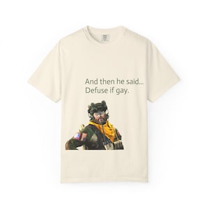 May include: Cream-colored t-shirt featuring a graphic of a soldier and the text "And then he said... Defuse if gay." The soldier is wearing a yellow scarf and a military helmet. The shirt is made of a soft material.