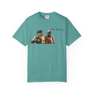 May include: Teal-colored t-shirt featuring a graphic of two men and the text "My Nigga". The men are wearing sunglasses and various clothing. The shirt is a short-sleeved, crew neck style.