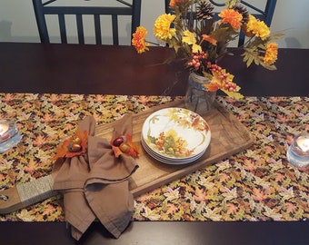 May include: A fall-themed table runner with a brown background and a pattern of colorful autumn leaves. The runner is set on a dark brown table and features a wooden serving tray with a stack of plates and two brown napkins with fall-themed napkin rings.