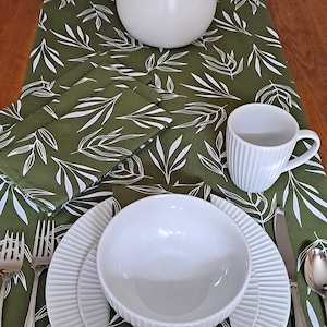 May include: A green table runner with a white leaf pattern. The runner is set with a white bowl, a white cup, and silverware.