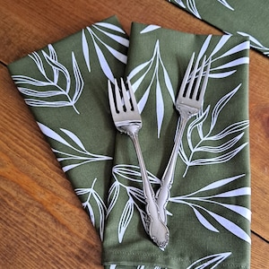 May include: Two green napkins with a white leaf pattern are folded together. Two silver forks are placed on top of the napkins.