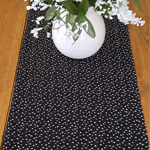 May include: Black and white table runner with a repeating pattern of small white triangles. A white vase with white flowers sits on top of the runner.