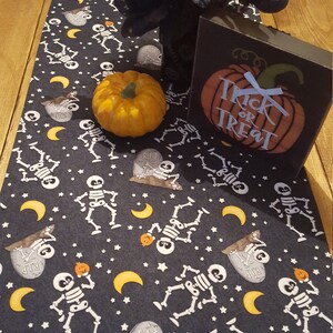 Peut inclure: Un chemin de table d'Halloween noir et blanc avec un motif répété de squelettes, d'étoiles, de lunes et de citrouilles. Le chemin de table est décoré d'une petite citrouille orange et d'une pancarte en bois qui dit "Trick or Treat".
