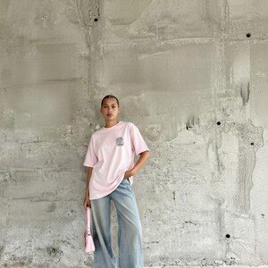 May include: A pale pink t-shirt with a small chest graphic, worn with light wash denim jeans. A matching pink handbag completes the outfit. The model is posing in front of a concrete wall.