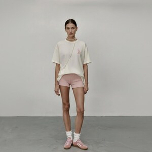 May include: A person wearing a cream t-shirt with a pink graphic, pink shorts, white socks, and pink trainers. The casual outfit is suitable for everyday wear. The background is a plain, light-coloured wall. The outfit is simple and casual.