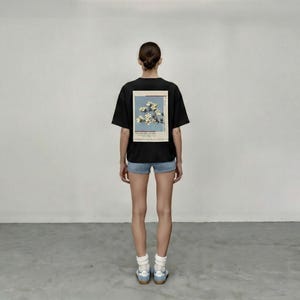 May include: A black t-shirt with a floral print on the back, worn with denim shorts and white socks. The graphic shows white flowers on a light blue backdrop. The person is wearing blue trainers.