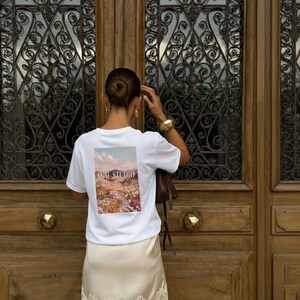 May include: White t-shirt with a colourful floral and landscape graphic on the back, featuring the text "SAAL STUDIO". The person is wearing a gold bracelet and earrings, and a cream-coloured skirt. The background is a wooden door.
