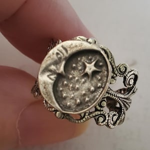 Puede incluir: Anillo plateado con un diseño de luna creciente y estrella dentro de un marco ovalado. El anillo tiene una banda ornamentada de estilo filigrana con detalles intrincados. La luna y las estrellas están en relieve, creando una superficie texturizada.