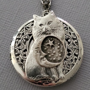 K&ouml;nnte beinhalten: Eine silberfarbene Medaillon-Halskette mit einem runden Anh&auml;nger, der einen filigranen Rand aufweist. In der Mitte befindet sich ein Relief einer Katze, die eine Mondsichel mit einem Stern und kleinen Punkten h&auml;lt, an einer passenden Gliederkette.