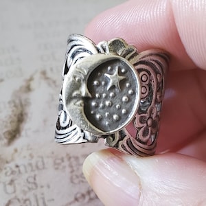Puede incluir: Anillo plateado con un diseño de luna creciente con cara, una estrella y pequeños puntos. El anillo tiene un diseño floral y de filigrana. El anillo está sostenido por una persona.