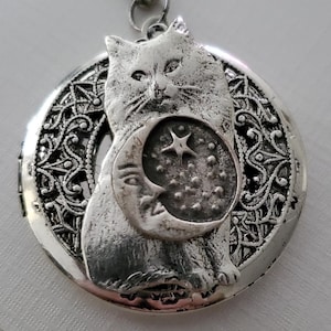 K&ouml;nnte beinhalten: Ein silberfarbener, runder Medaillon-Anh&auml;nger an einer passenden Kette. Das Medaillon zeigt ein erhabenes Relief einer Katze, die einen Halbmond mit einem Stern vor einem texturierten Hintergrund h&auml;lt. Der Rand hat ein filigranes Muster.