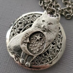 K&ouml;nnte beinhalten: Ein silberfarbener, runder Medaillon-Anh&auml;nger an einer passenden Kette. Das Medaillon zeigt ein erhabenes Relief einer Katze vor einem Halbmond- und Sternmotiv, eingebettet in einen Hintergrund aus filigranen, metallischen Schn&ouml;rkeln.