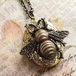 May include: A gold-toned locket necklace featuring a detailed bee design. The locket is oval-shaped and has a floral pattern around the bee.