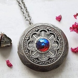 May include: A silver locket necklace with a detailed filigree design. The locket features a round, red and blue iridescent stone in the center.