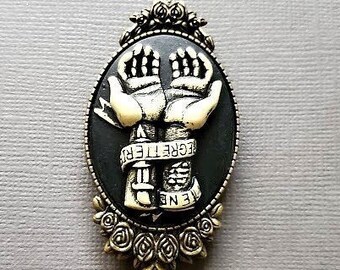 May include: An oval-shaped pendant with a black background featuring two hands reaching upwards. The hands are cream-colored, and the pendant is framed with a decorative silver-toned border and floral accents. The word "REGRETTER" is visible.