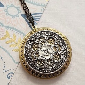 Puede incluir: Un collar con un medallón de plata y oro con un diseño floral detallado. El medallón tiene un patrón de filigrana y una pequeña flor en el centro.