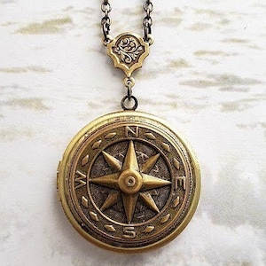 Puede incluir: Un collar con un medallón de brújula de oro y una cadena negra. El medallón presenta una rosa de los vientos con las letras N, S, E y W alrededor del borde.