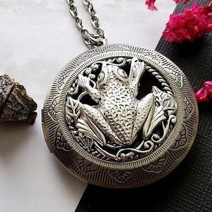May include: Silver necklace with a round locket featuring an ornate design of a frog surrounded by leaves. The locket is open and the inside is visible.
