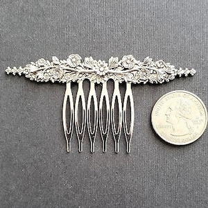 May include: A silver hair comb with a floral design. The comb has seven teeth and is about the size of a US quarter.